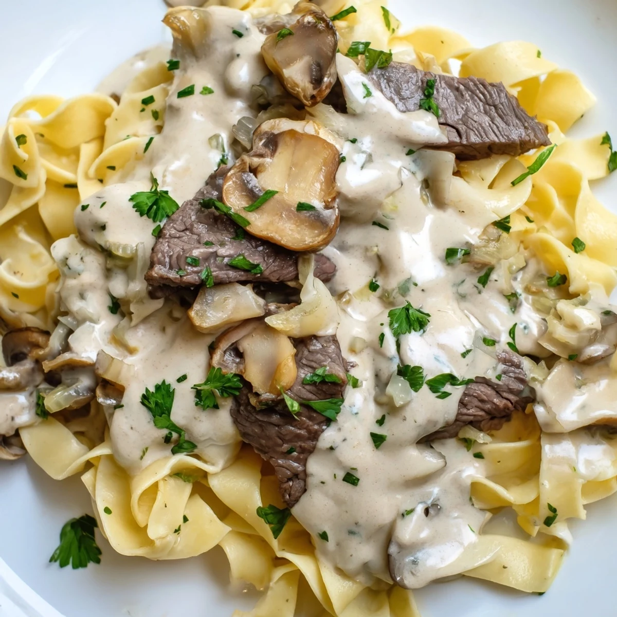 Tender beef strips and cremini mushrooms in a creamy sauce are ladled over steaming egg noodles, garnished with fresh parsley.