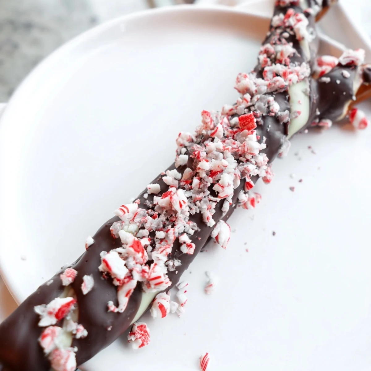 A hand holding a dark chocolate peppermint bark pretzel with white chocolate drizzle and crushed peppermint.