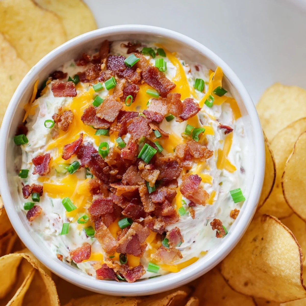 Homemade Loaded Baked Potato Dip in a white bowl, garnished with green onions and cheddar, surrounded by golden potato chips.