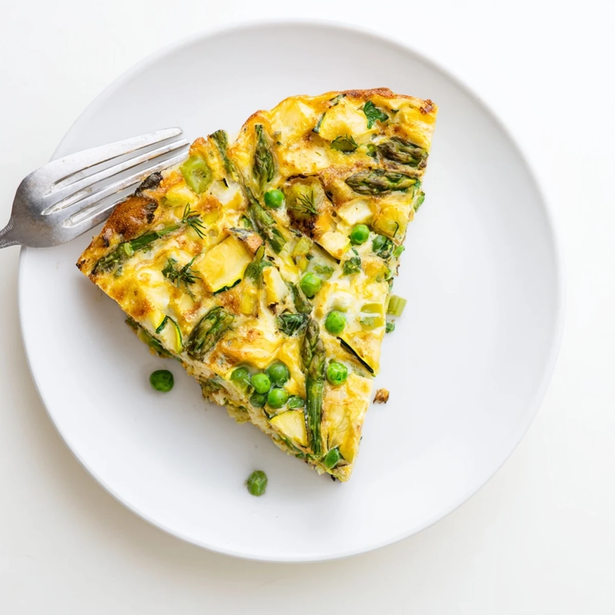 The golden-brown Spring Vegetable Frittata with Herbs, featuring asparagus, spinach, and peas, rests on a white plate.  